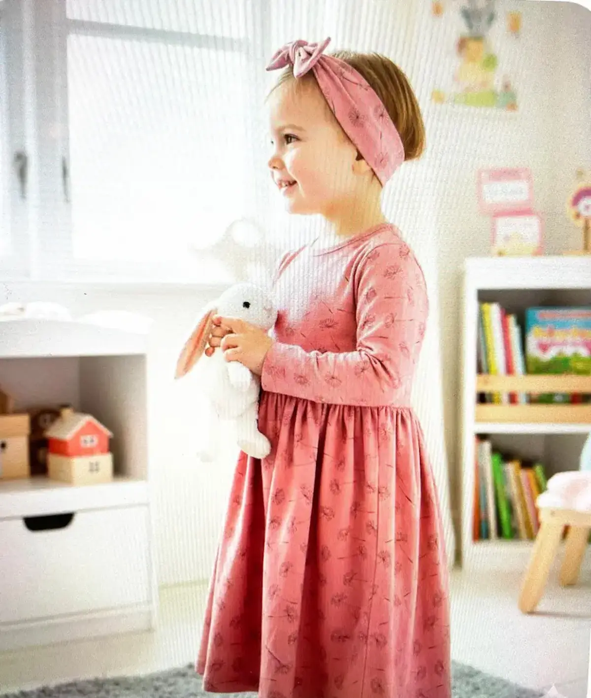 Weicher Baumwolljersey Stoff mit Pusteblumen-Motiv als Meterware, genäht als altrosafarbenes Kinderkleid mit passendem Haarband in einem hellen Kinderzimmer.