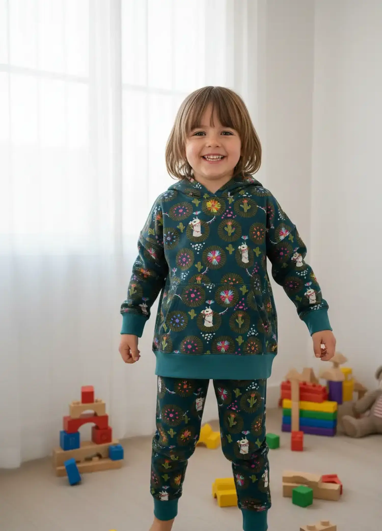 Tragebeispiel für den elastischen French Terry Stoff in Petrol mit Lama- und Kaktus-Motiv: Ein fröhliches Kind trägt einen selbstgenähten Kapuzenpullover und eine passende Jogginghose im hellen Kinderzimmer beim Spielen mit Holzbausteinen.