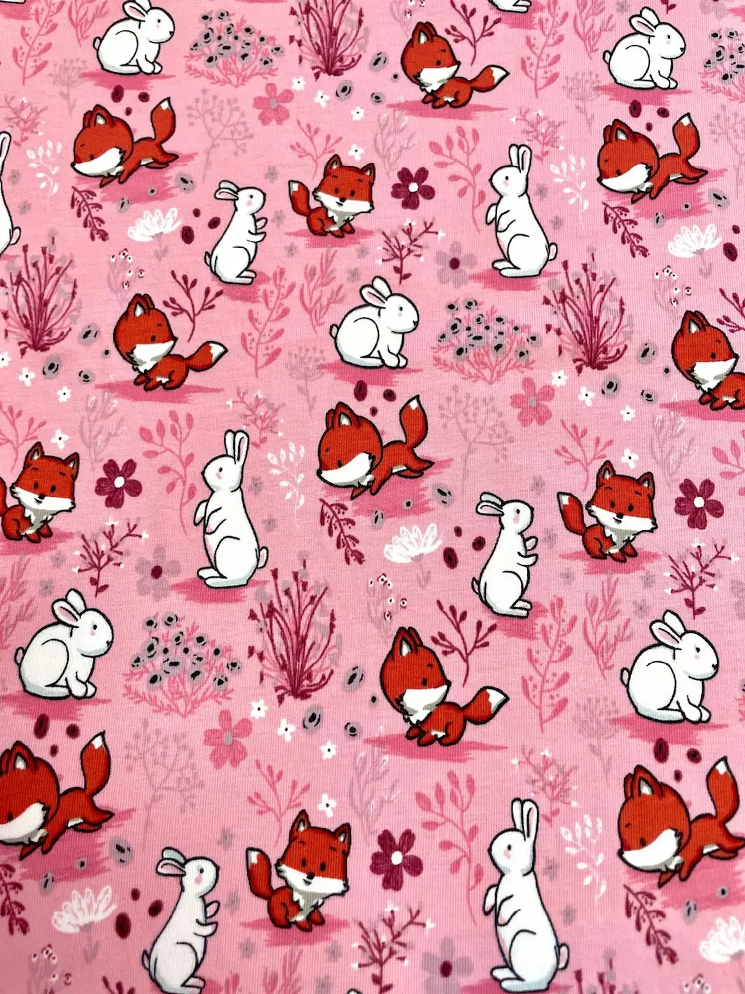 Niedlicher Baumwolljersey in Rosa mit kleinen Füchsen, weißen Hasen und floralen Elementen wie Blumen und Gräsern, perfekt für Baby- und Kinderkleidung.