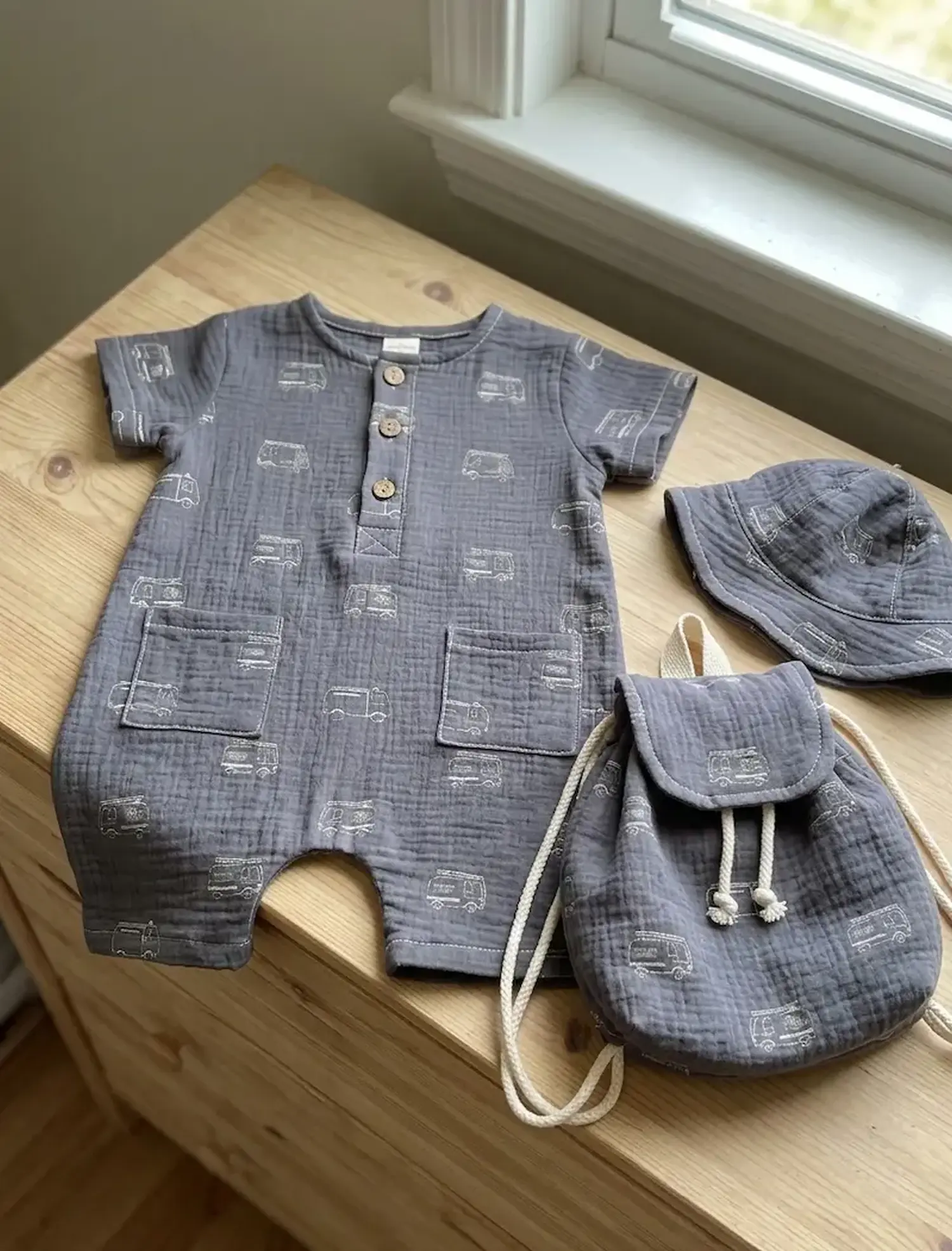 Baby-Set aus grauem Musselin mit weißem Feuerwehrauto-Aufdruck, bestehend aus einem kurzen Einteiler (Romper), passendem Sonnenhut und einem kleinen Stoff-Rucksack auf einer Holzoberfläche.