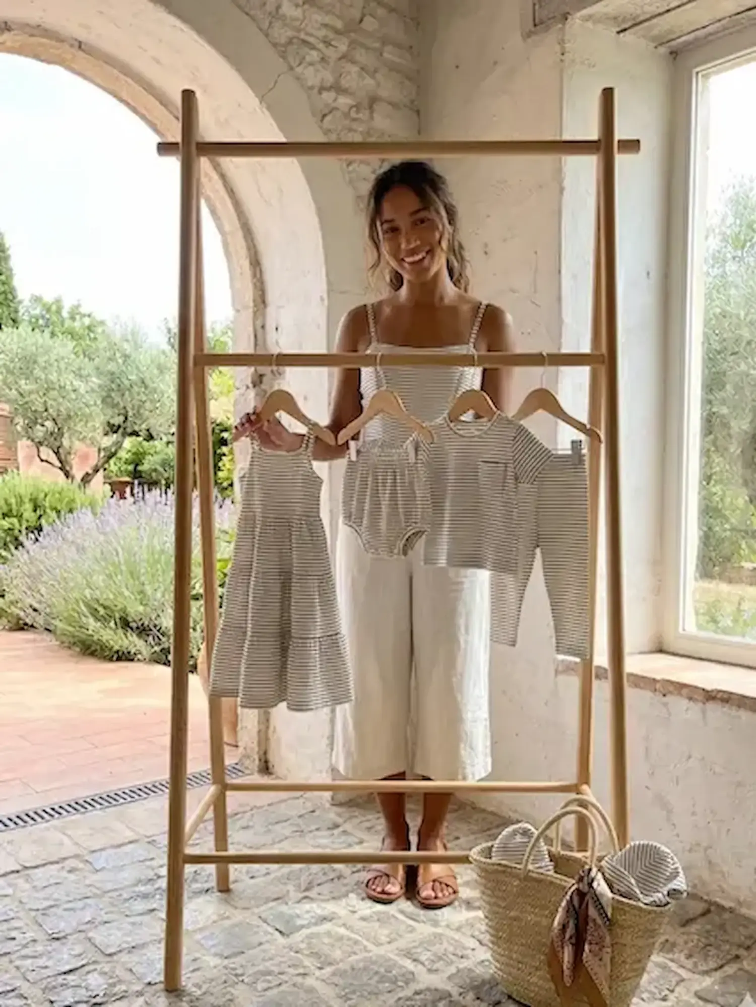 Eine lächelnde Frau präsentiert eine Auswahl an Kinderbekleidung aus gestreiftem Jersey-Stoff (Kleid, Shirt, Hose) an einem Holzständer vor einer mediterranen Steinkulisse