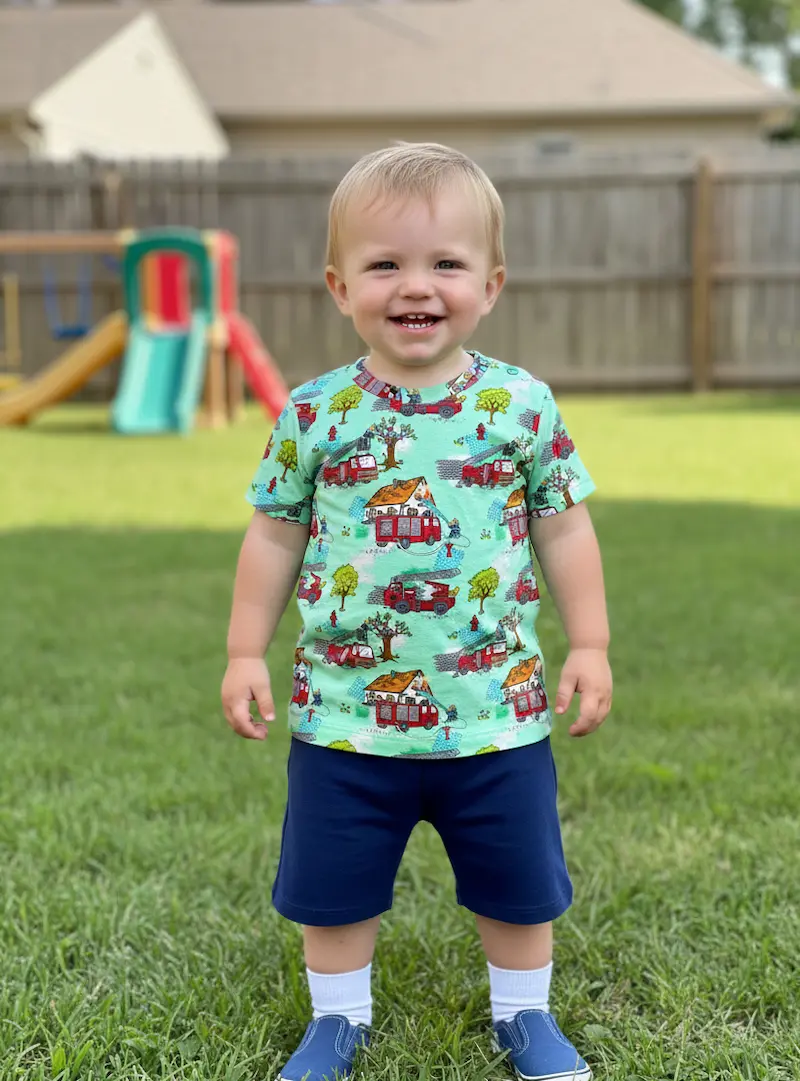 Kleinkind im Garten trägt ein selbstgenähtes T-Shirt aus hellgrünem Jersey-Stoff mit bunten Feuerwehrauto-Motiven.