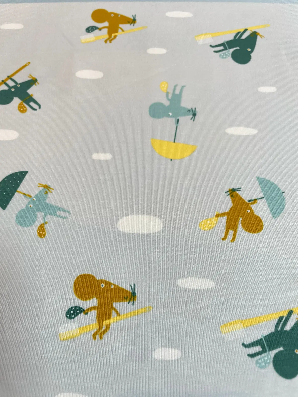 Ein flach liegender Baumwolljersey-Stoff als Meterware in hellem Graublau, bedruckt mit einem niedlichen Kindermuster aus kleinen ockerfarbenen und grünen Mäusen, die Zahnbürsten tragen oder mit bunten Regenschirmen an weißen Wolken vorbeifliegen.