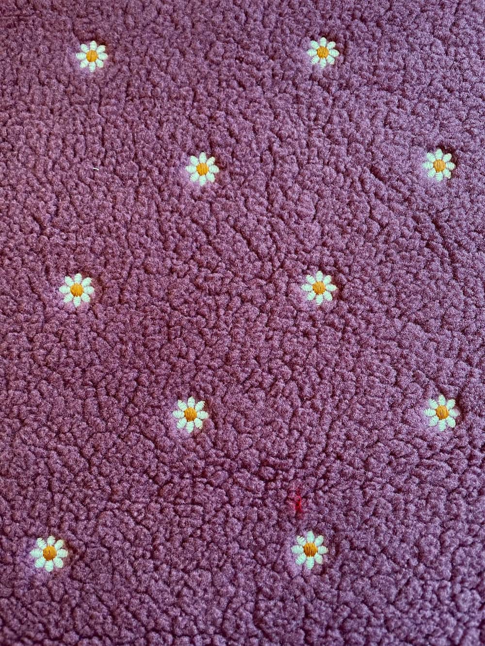 Detailaufnahme von weichem Teddyplüsch in Beere-Mauve mit feinen Gänseblümchen-Stickereien als Meterware.
