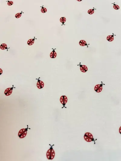 Heller, weißer Jersey-Stoff mit einem niedlichen Streumuster aus kleinen roten Marienkäfern. Die Käfer sind detailliert mit schwarzen Punkten gezeichnet und gleichmäßig auf dem dehnbaren Baumwolljersey verteilt. Perfekt für Baby- und Kinderkleidung.
