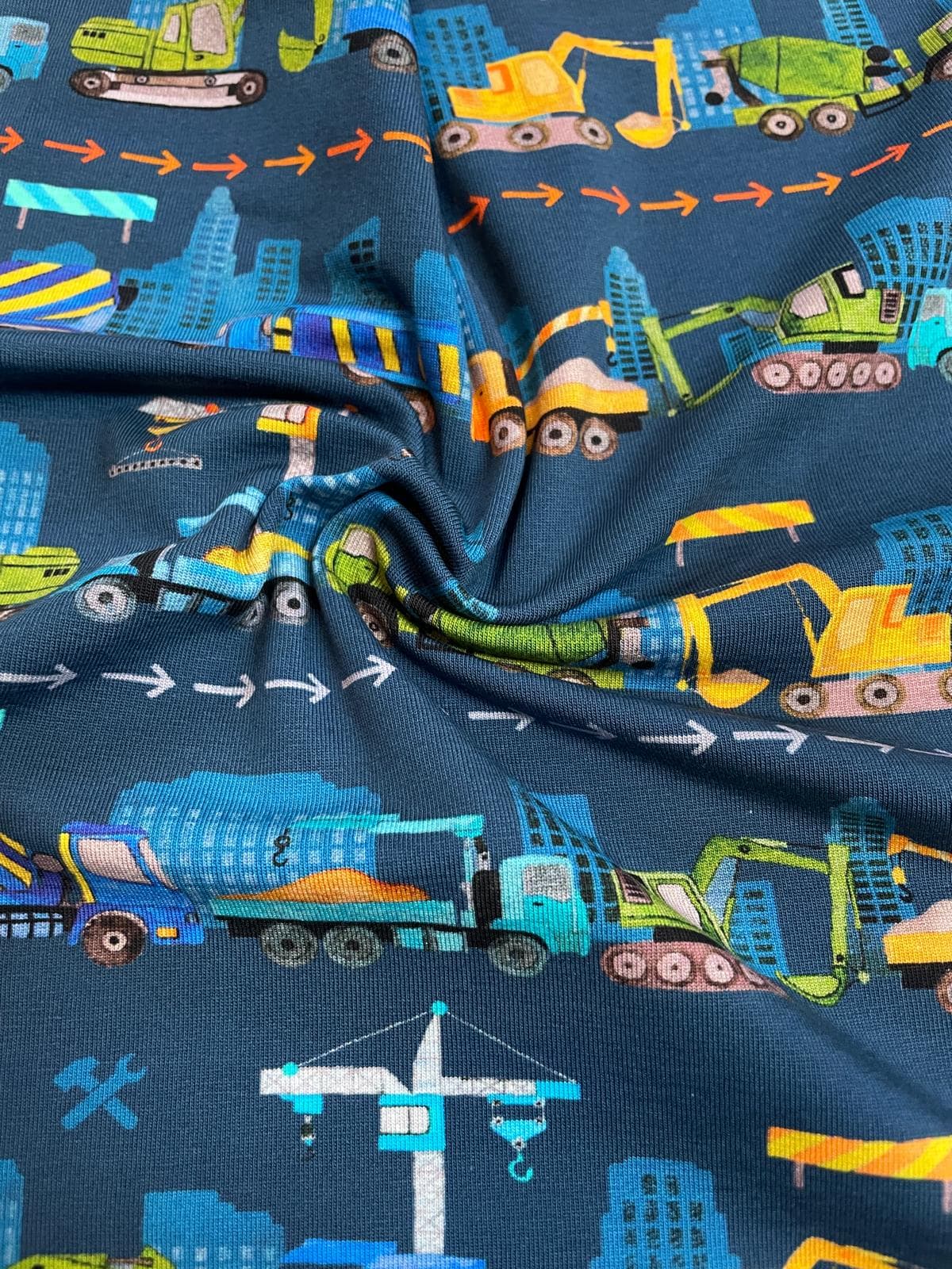Ein kunstvoll drapierter, dunkelblauer Jerseystoff als Meterware, der das weiche Material zeigt und mit bunten Baggern, Kränen und Betonmischern vor einer städtischen Skyline bedruckt ist.