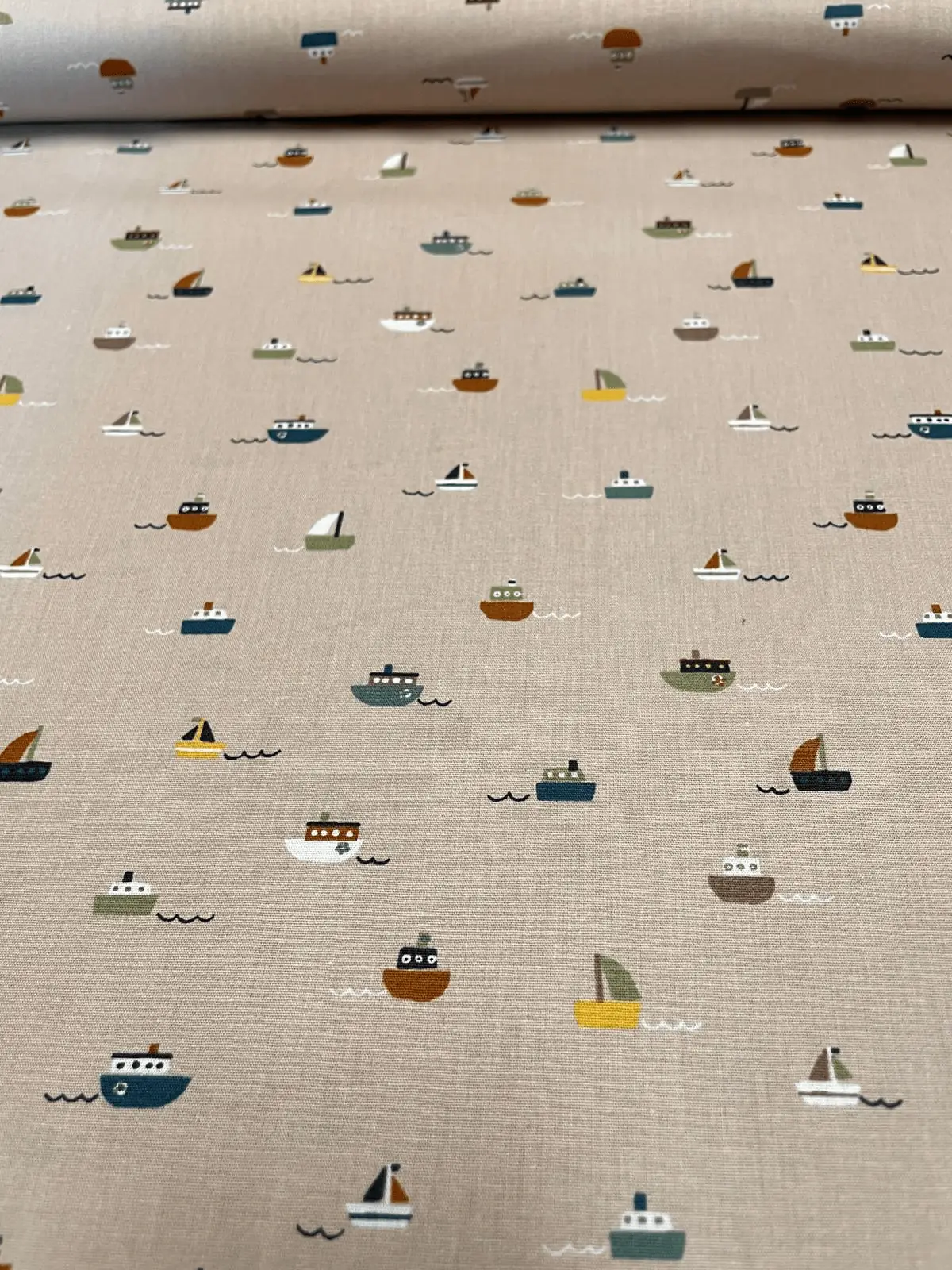 Draufsicht auf hellbraune bzw. beige Bio-Baumwoll-Popeline als Meterware mit einem feinen, minimalistischen Muster aus kleinen bunten Segelbooten und Dampfern auf minimalistischen Wellen