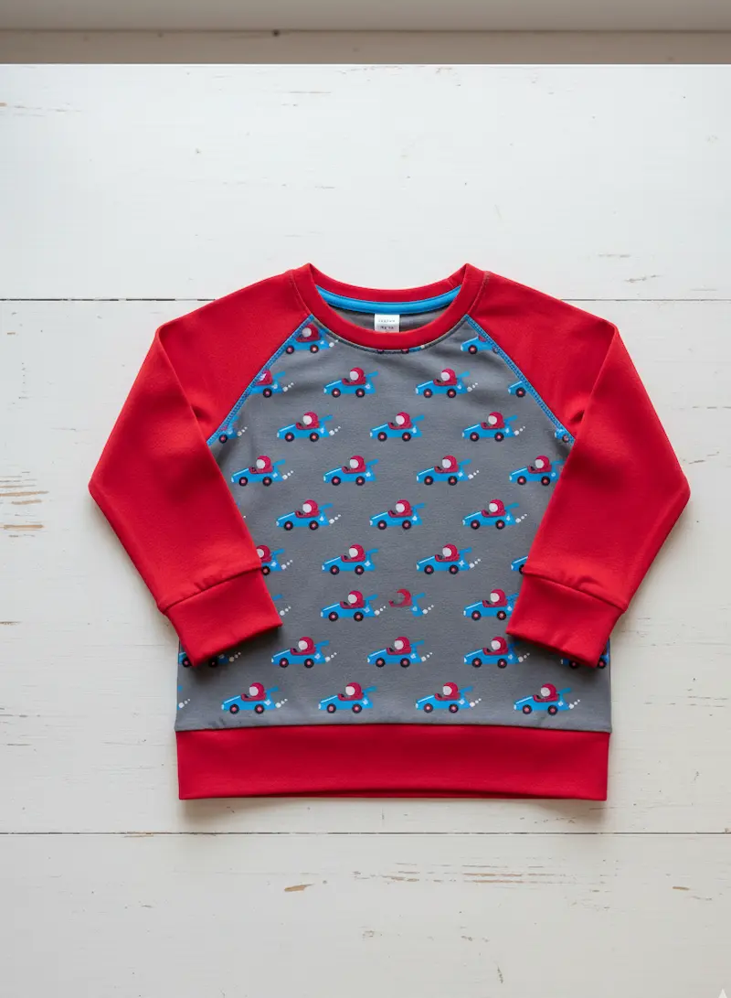 Ein flach liegender Kinder-Raglanpullover auf einer weißen Holzoberfläche. Das Vorderteil besteht aus grauem Baumwolljersey mit kleinen blauen Rennautos, kombiniert mit roten Ärmeln und roten Bündchen aus Jersey oder Sweat.