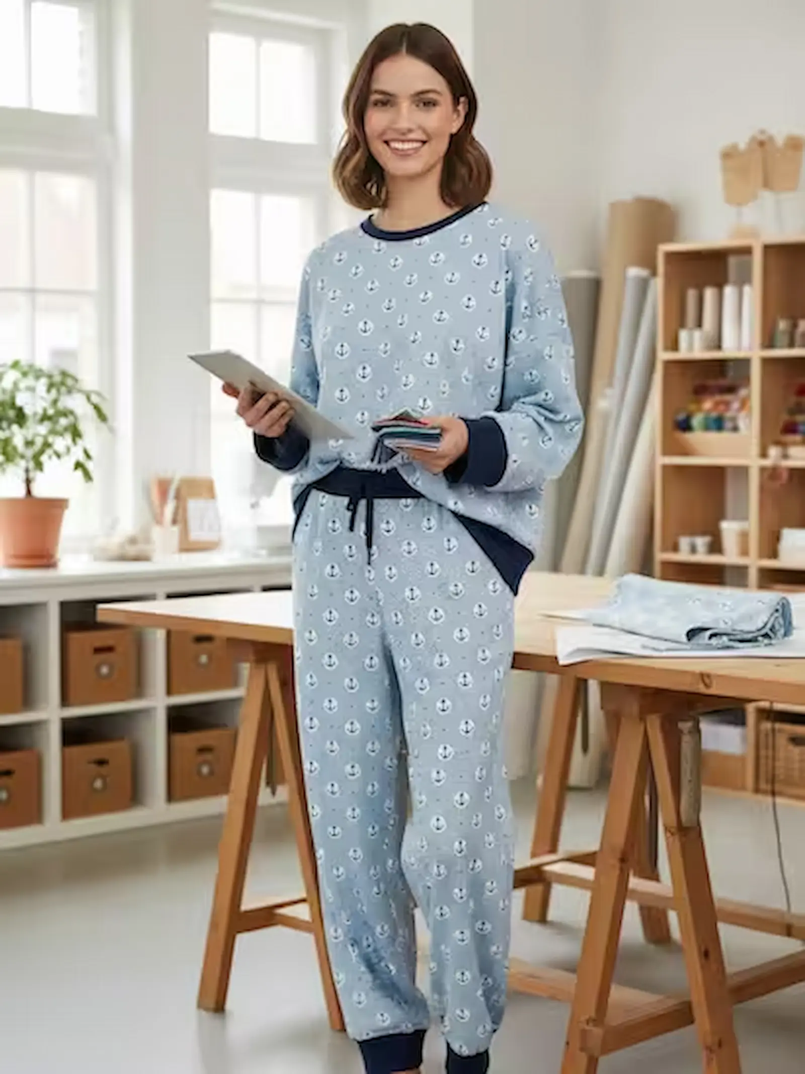 Eine lächelnde Frau in einem selbstgenähten, gemütlichen Zweiteiler aus hellblauem Baumwolljersey mit weiß-dunkelblauen Ankermotiven. Sie steht in einem hellen, freundlichen Nähzimmer an einem Zuschneidetisch aus Holz, hält ein Tablet und Stoffmuster in d