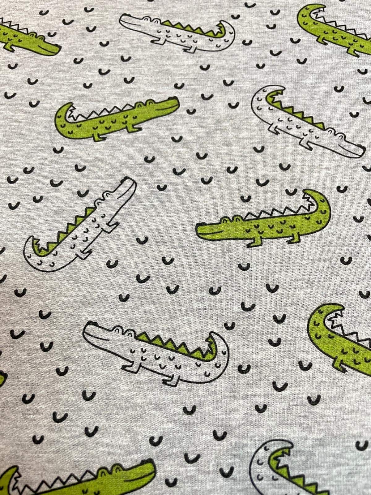 Kuscheliger Alpenfleece-Stoff in Grau-Melange mit Krokodil-Print. Niedliches Reptilien-Muster mit grünen Krokodilen und kleinen Wellen, ideal für warme Kinderkleidung und Decken.