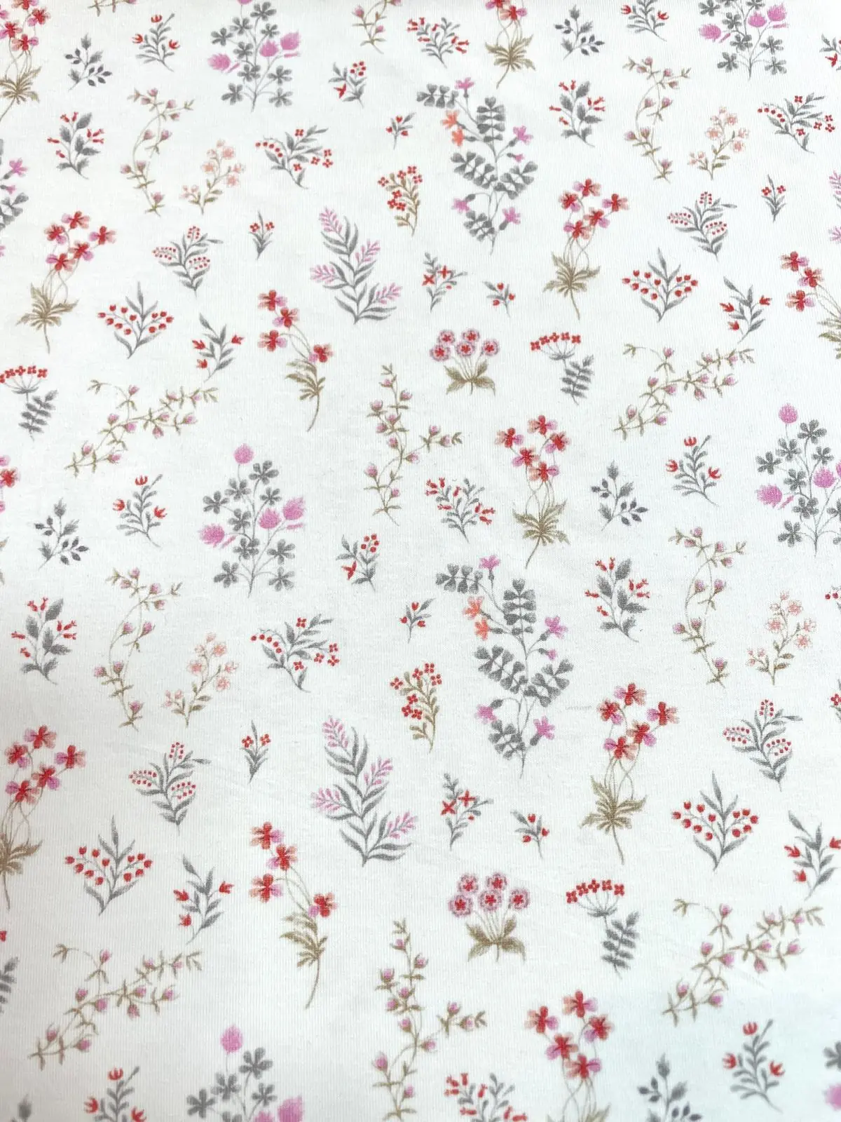 Weißer Jersey-Stoff mit einem feinen, floralen Muster aus kleinen bunten Wiesenblumen in Rosa, Rot und Grau.