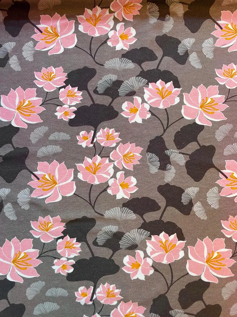 Elastischer Jerseystoff als Meterware mit floralem Muster aus rosa Lotusblüten und grau-braunen Ginkgoblättern.