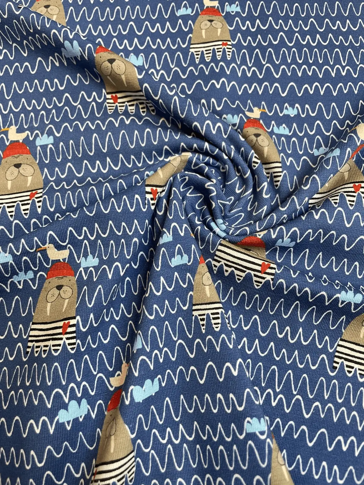 Ein weich drapierter Baumwolljersey-Stoff als Meterware in Dunkelblau mit weißen Zickzack-Wellenlinien. Im Faltenwurf sind die aufgedruckten Motive von lustigen Walrossen mit roten Mützen, kleinen Möwen und hellblauen Wolken zu sehen.