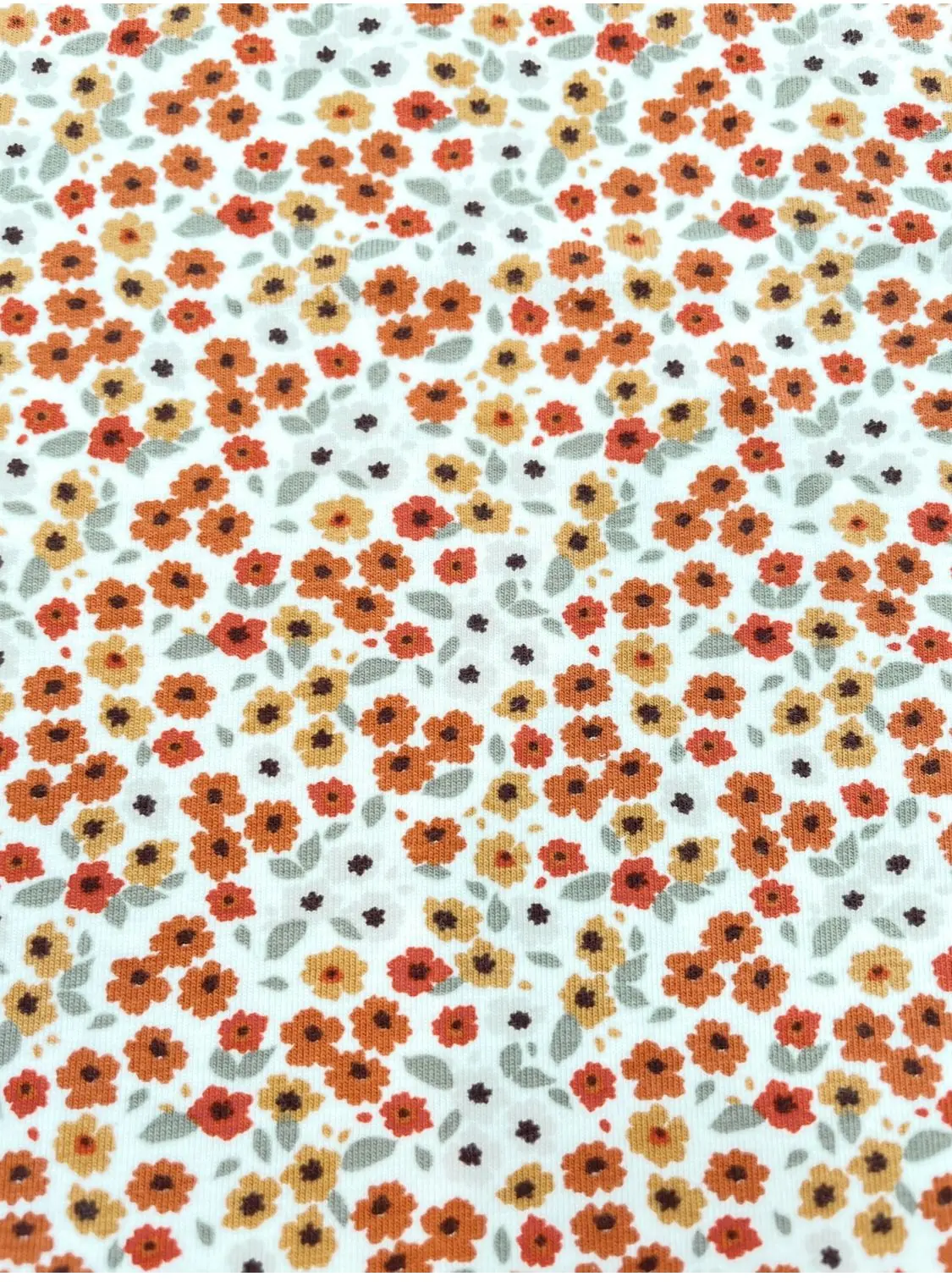 Elastischer Jersey-Stoff in Weiß mit einem dichten Muster aus kleinen Streublümchen in Orange, Ockergelb und Braun mit hellgrünen Blättern.