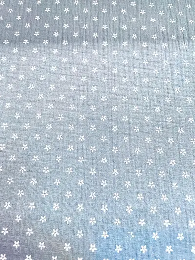 Flächenansicht von taubenblauem Musselin-Stoff mit einem Allover-Print aus kleinen weißen Blüten. Die Aufnahme zeigt die regelmäßige Musterverteilung und die charakteristische Crinkle-Struktur der Double-Gauze Meterware.