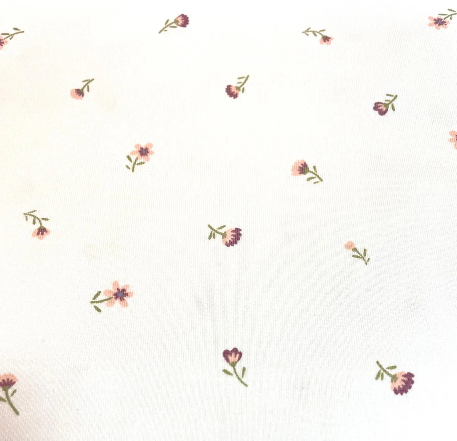Weißer Jersey-Stoff mit einem feinen Muster aus kleinen, zarten Streublumen in Rosa, gedecktem Lila und grünen Stielen.