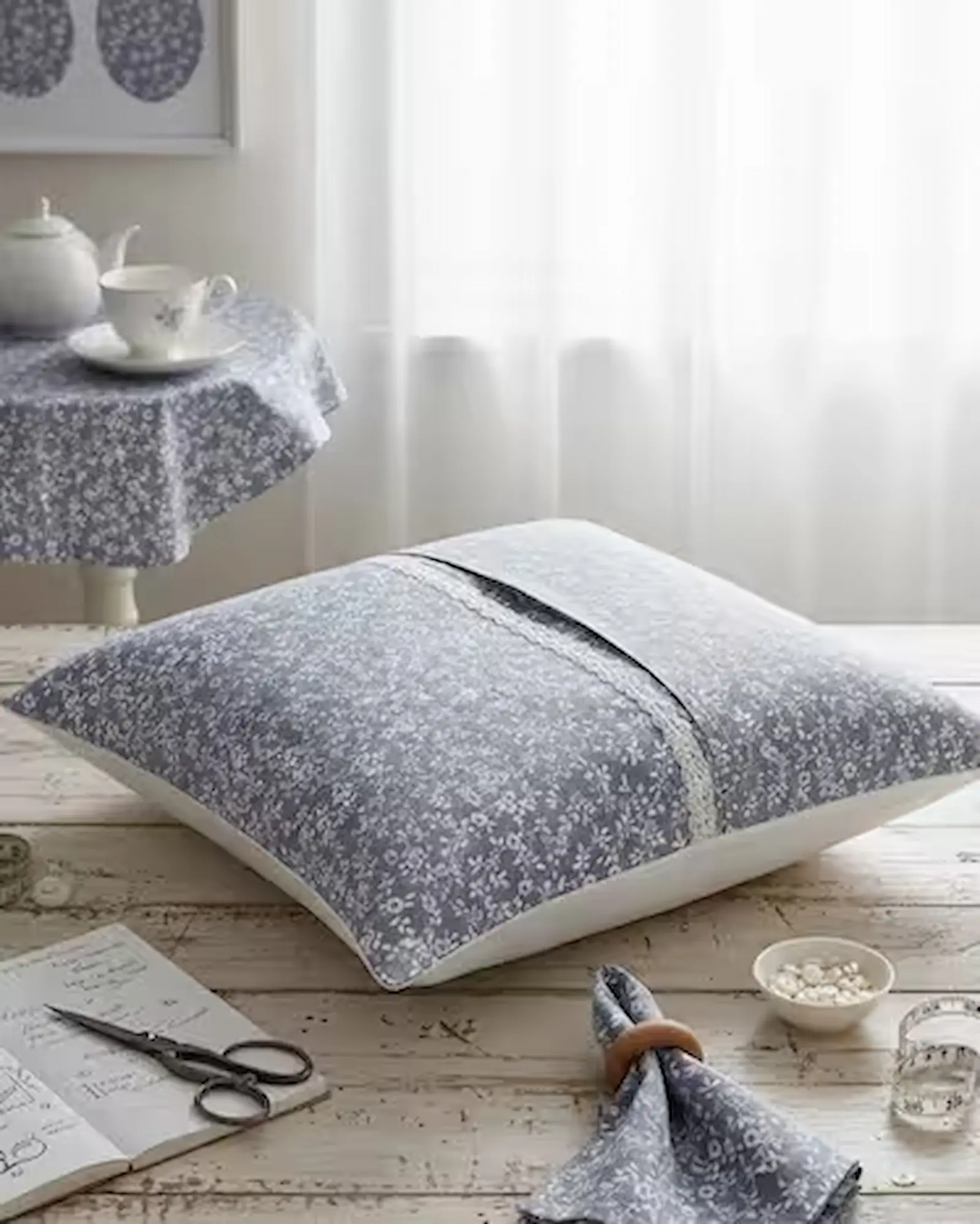 Ein selbstgenähtes, großes Dekokissen aus grauer Baumwoll-Popeline mit einem feinen, weißen Blumenmuster (Streublumen). Das Kissen ist mit einem weißen Spitzenband verziert und liegt auf einem rustikalen Holztisch. Im Hintergrund ist ein runder Tisch mit