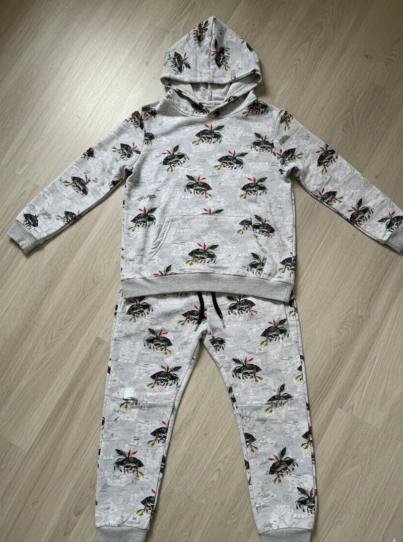 Ein selbstgenähtes Set aus Hoodie und passender Jogginghose für Kinder, flach liegend auf hellem Boden. Das Set besteht aus hellgrauem Baumwolljersey mit einem Allover-Print aus Piraten-Totenköpfen und Schatzkarten-Motiven sowie hellgrauen Bündchen.
