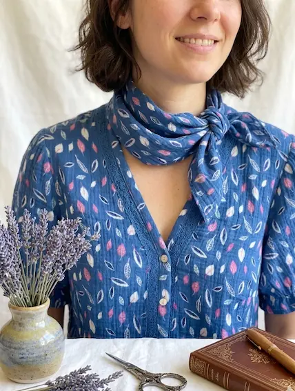 Eine lächelnde Frau trägt eine Bluse und ein passendes Halstuch aus blauem Musselin mit Blättermuster; auf dem Tisch liegen Lavendel, eine Schere und ein Buch.