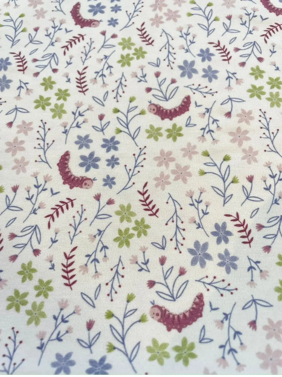 Weißer Jerseystoff bedruckt mit niedlichen lila Raupen und einem zarten Blumenmuster in Pastellfarben wie Flieder, Rosa und Hellgrün, ideal für Babykleidung.