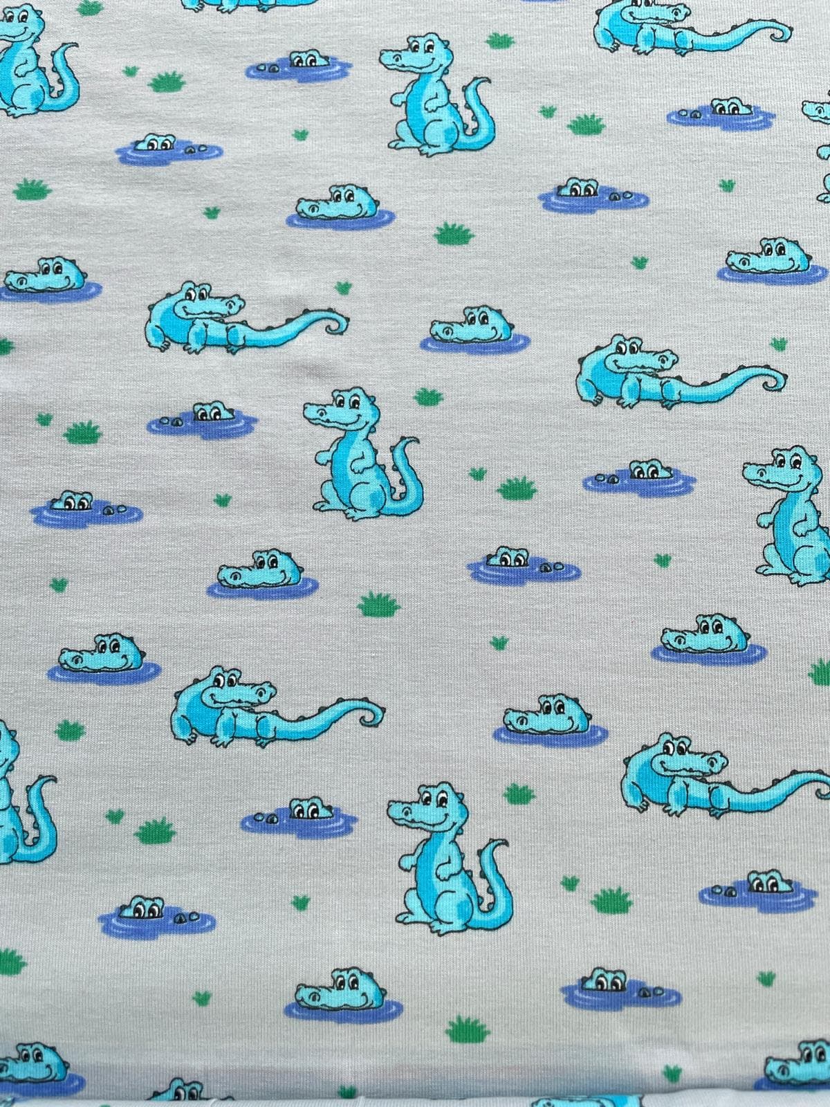 Hochwertiger Baumwolljersey als Meterware in Hellgrau Melange, bedruckt mit einem niedlichen Comic-Muster aus hellblauen Krokodilen. Die Alligatoren sitzen auf dem Land, schwimmen im Wasser oder gucken aus kleinen Sumpflöchern hervor, verziert mit kleinen