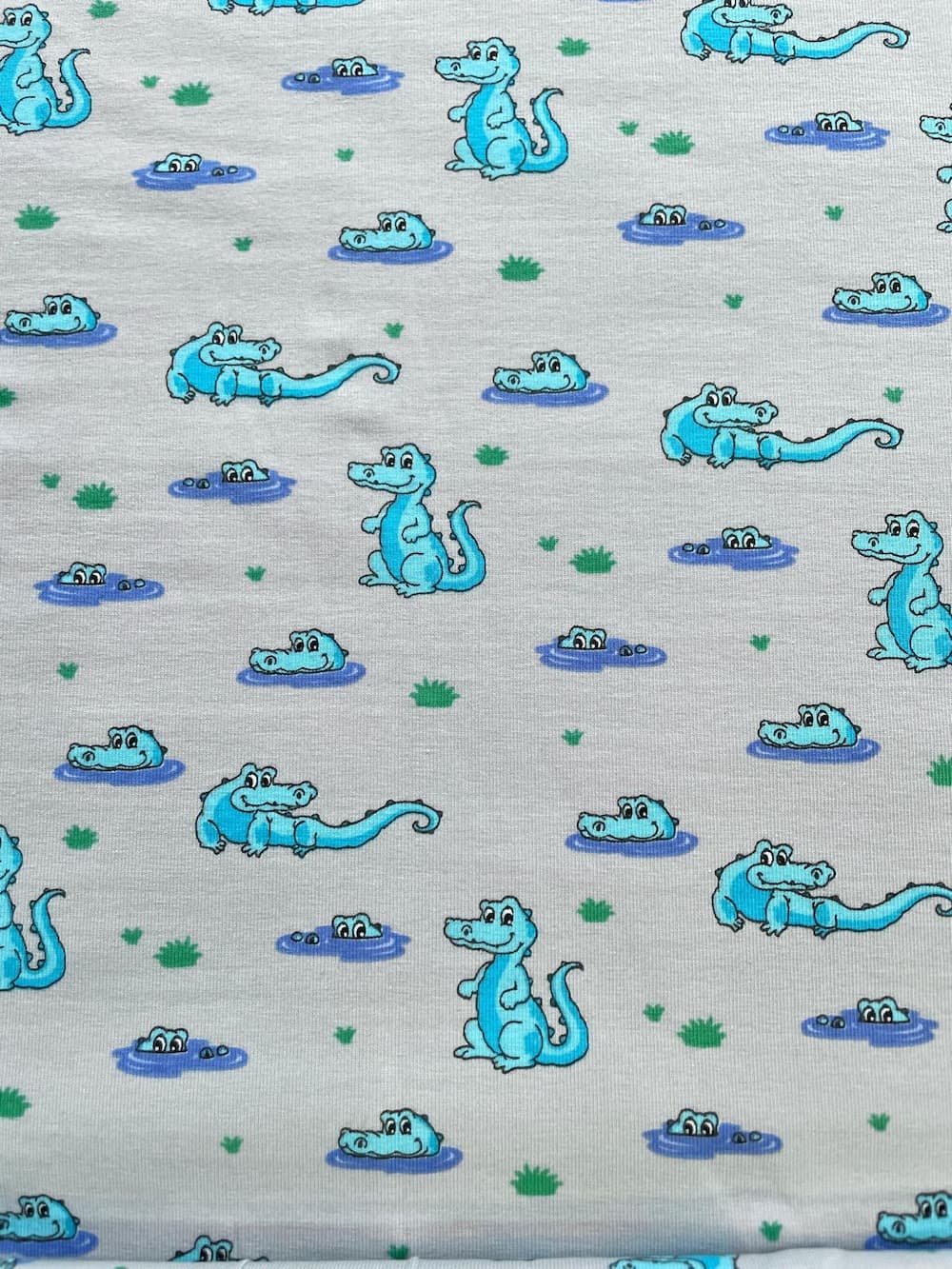 Hochwertiger Baumwolljersey als Meterware in Hellgrau Melange, bedruckt mit einem niedlichen Comic-Muster aus hellblauen Krokodilen. Die Alligatoren sitzen auf dem Land, schwimmen im Wasser oder gucken aus kleinen Sumpflöchern hervor, verziert mit kleinen