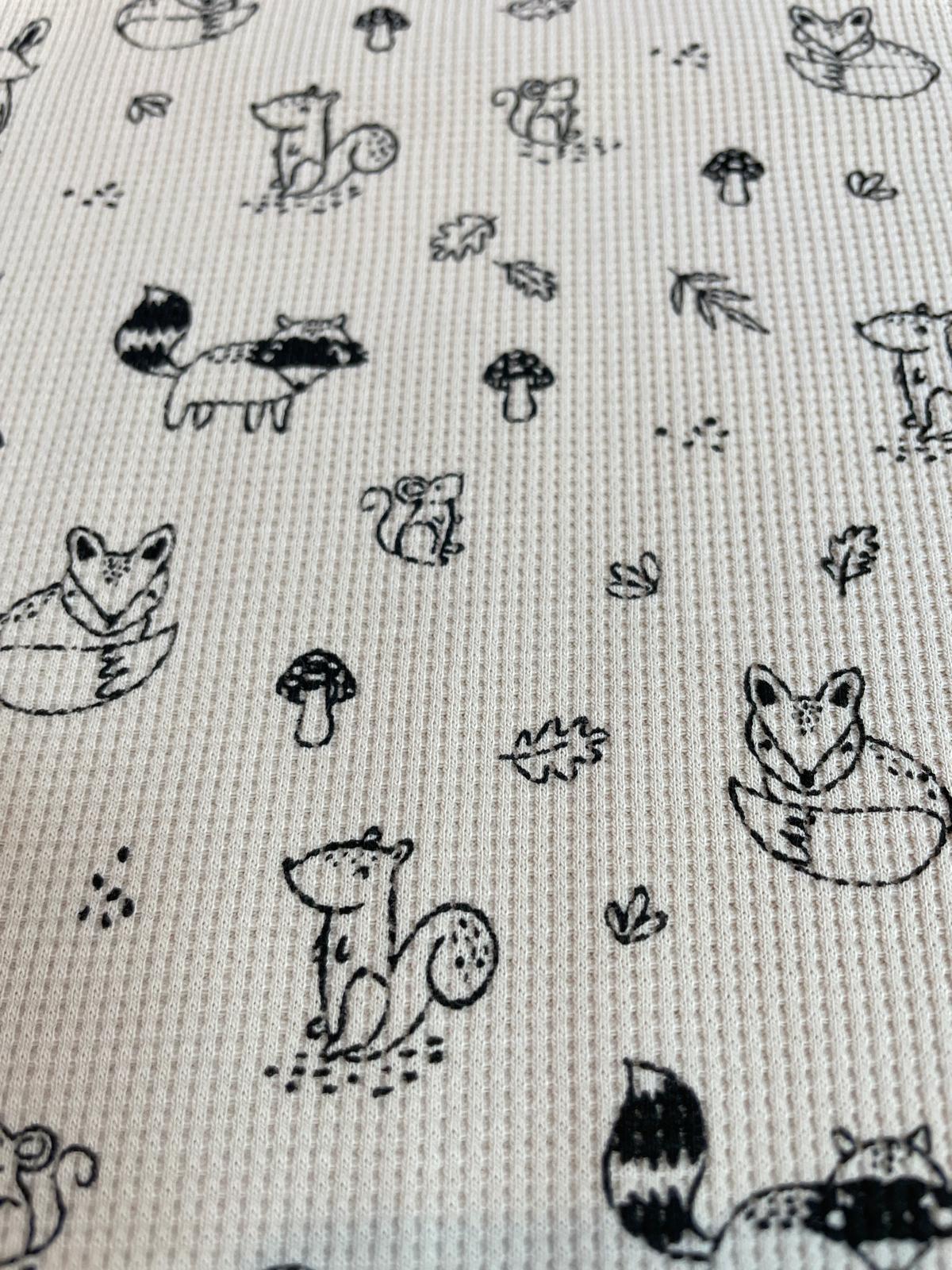 Strukturierter Waffelstrick bzw. Waffeljersey als Meterware in Beige mit einem feinen, schwarz-weißen Muster aus Waldtieren, Blättern und Pilzen.