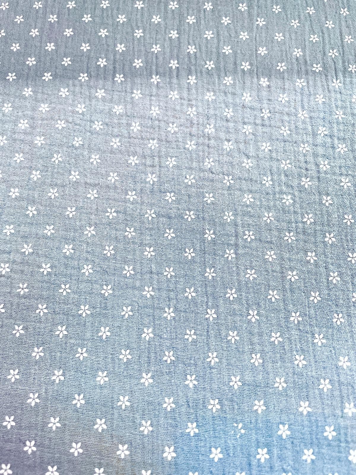 Flächenansicht von taubenblauem Musselin-Stoff mit einem Allover-Print aus kleinen weißen Blüten. Die Aufnahme zeigt die regelmäßige Musterverteilung und die charakteristische Crinkle-Struktur der Double-Gauze Meterware.