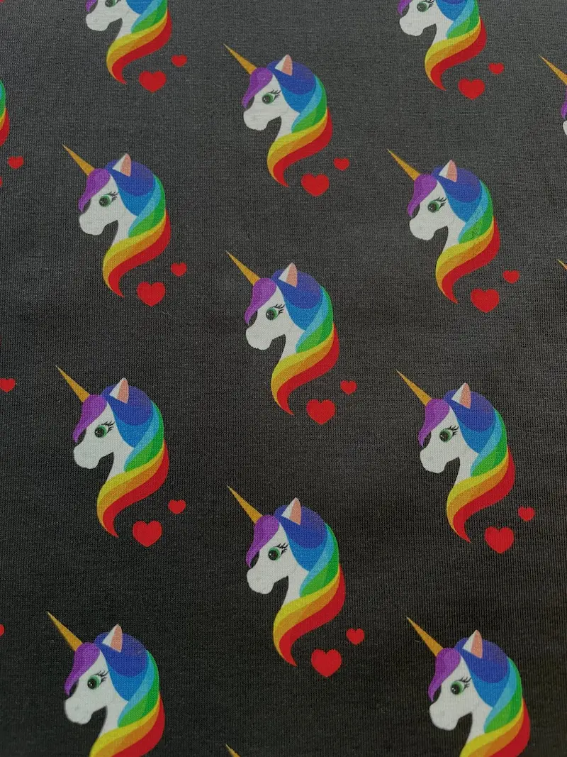 Hochwertiger Baumwolljersey als Meterware in Anthrazit-Dunkelgrau, bedruckt mit einem fortlaufenden Muster aus weißen Einhornköpfen, farbenfroher Regenbogen-Mähne und kleinen roten Herzen.