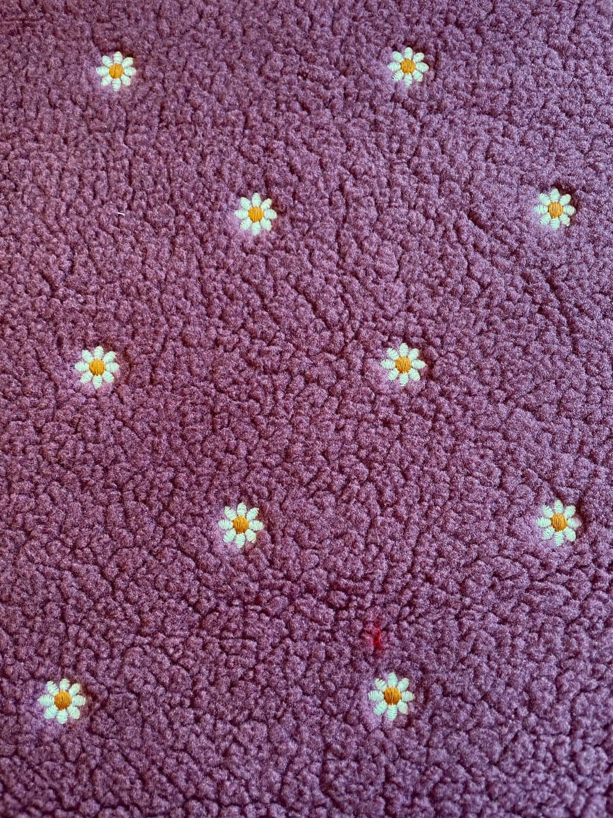 Detailaufnahme von weichem Teddyplüsch in Beere-Mauve mit feinen Gänseblümchen-Stickereien als Meterware.