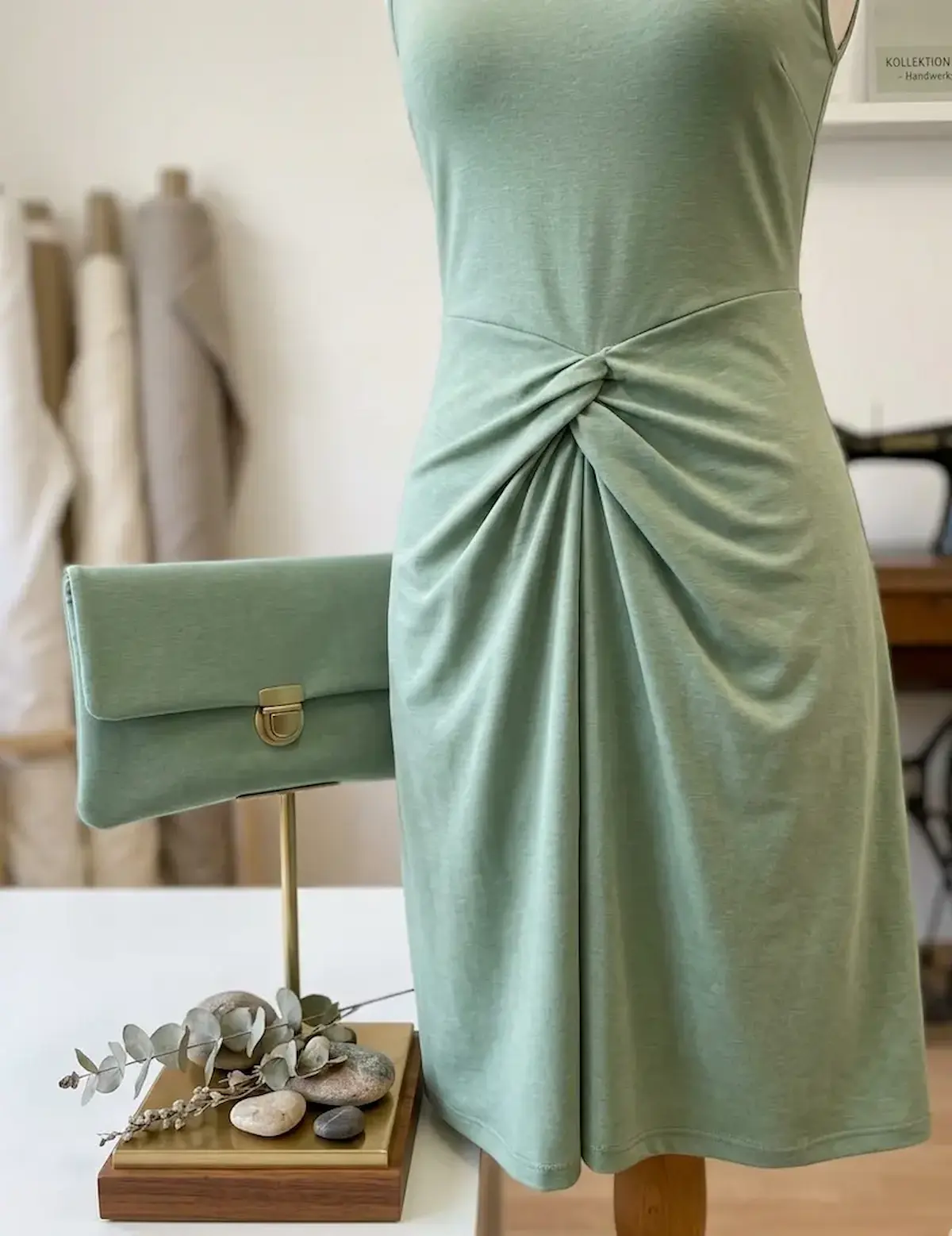 Detailansicht eines Kleides mit Knotendetail und einer passenden Tasche aus salbeigrünem Baumwolljersey Uni auf einer Schneiderpuppe.