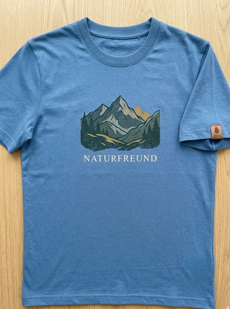Mittelblaues T-Shirt aus Baumwolljersey Uni Meterware mit Naturfreund-Bergmotiv-Aufdruck und Leder-Label auf Holzgrund.