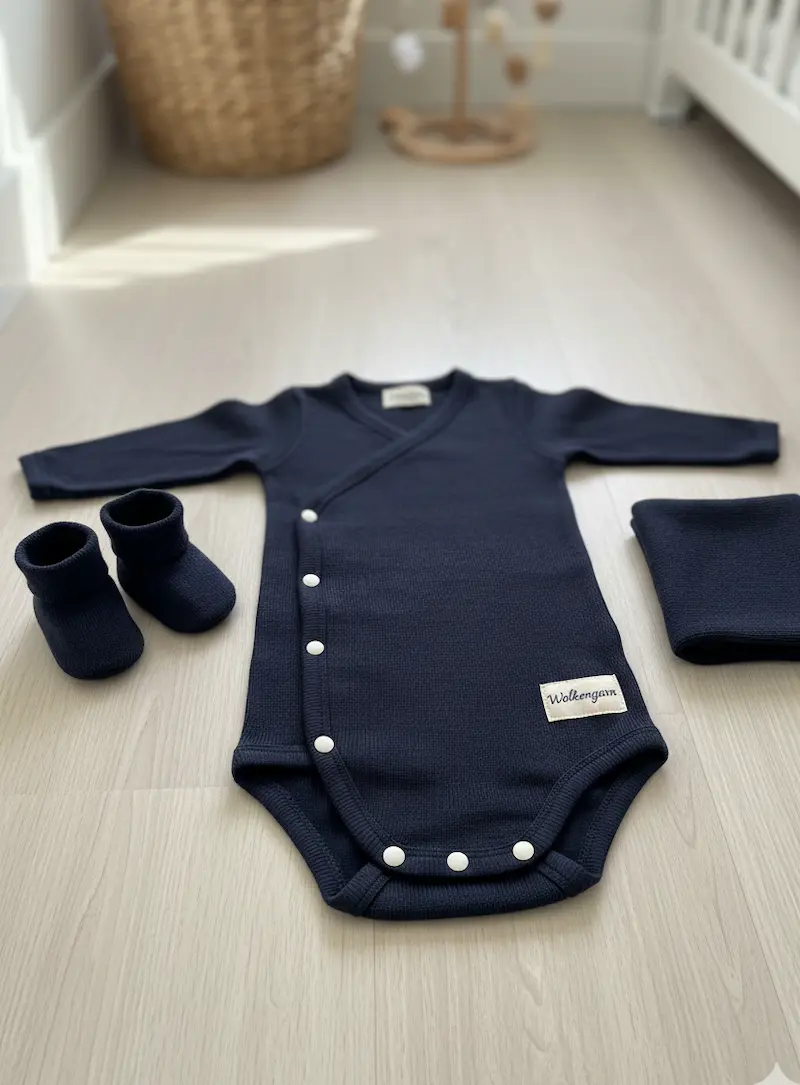 Dunkelblauer Baby-Wickelbody mit Druckknöpfen, passenden Booties und gefaltetem Feinstrick-Stoff aus Baumwolle mit Wolkengarn-Label auf Holzboden.