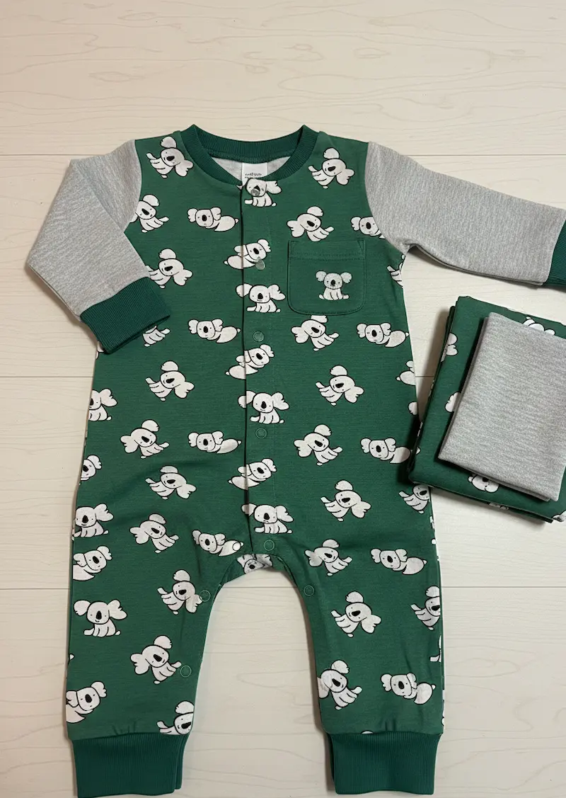 Selbstgenähtes Baby-Set bestehend aus einem dunkelgrünen Koala-Strampler und passend kombinierten grauen Jersey-Stoffen auf hellem Hintergrund.