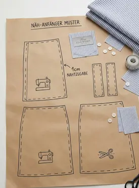 Schnittmuster für Nähanfänger auf braunem Papier mit 1cm Nahtzugabe neben blau-weiß gestreiftem Baumwollstoff und Nähzubehör