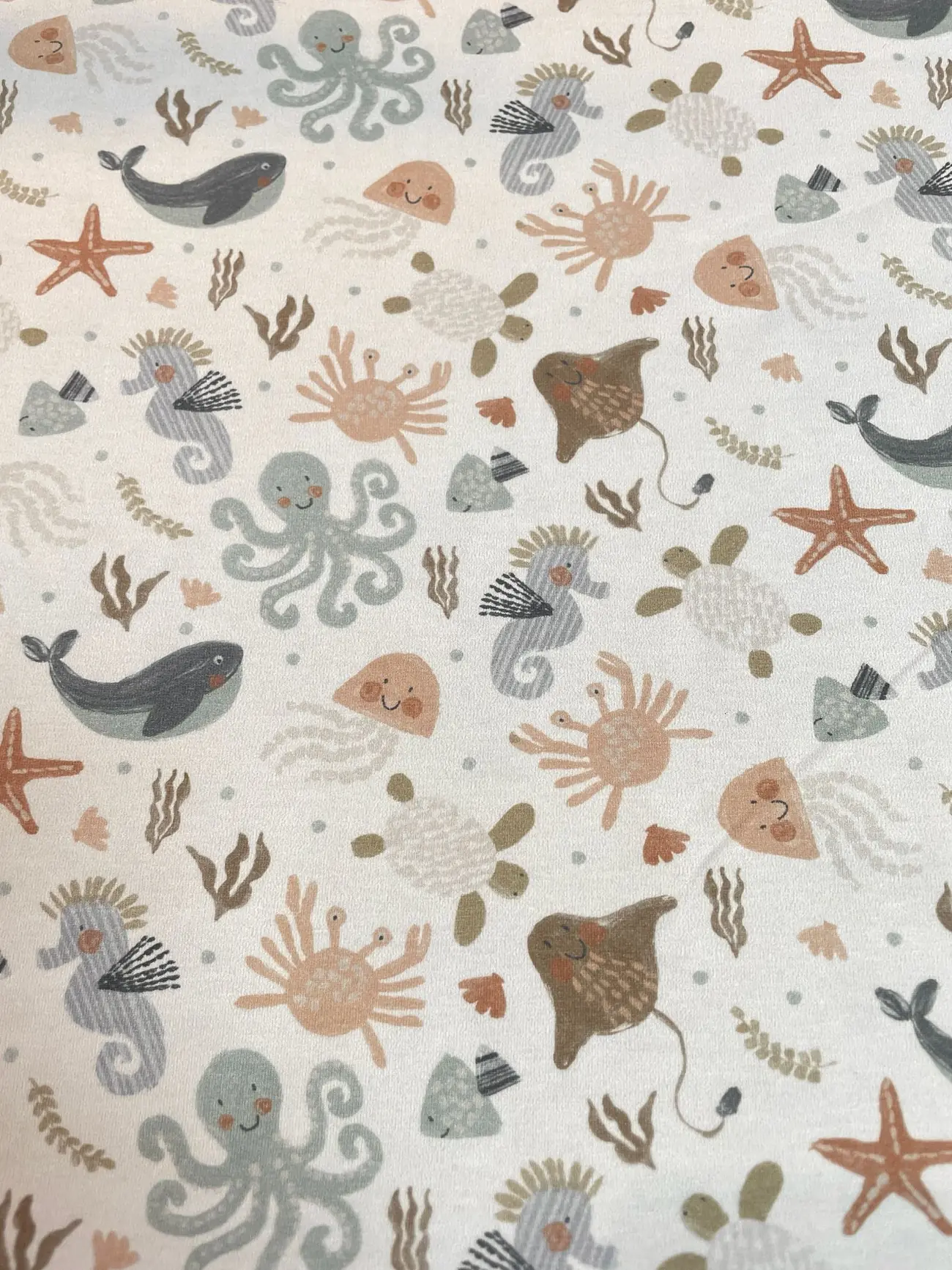 Cremeweißer Baumwolljersey-Stoff als Meterware, flach fotografiert und mit einem freundlichen Allover-Muster aus gezeichneten Ozean-Tieren wie Walen, Kraken, Seepferdchen, Rochen, Krabben, Quallen und Seesternen bedruckt.