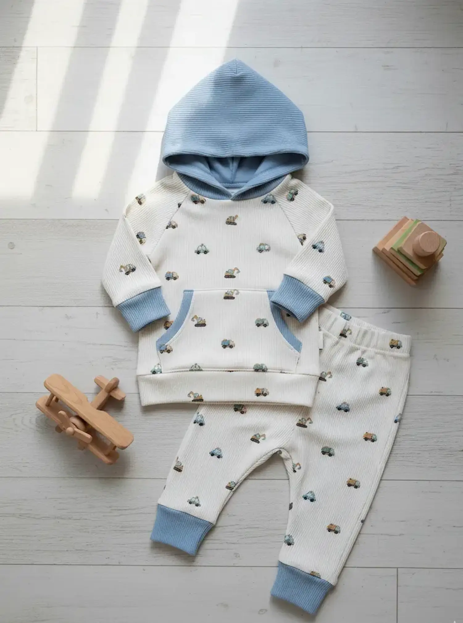 Ein genähtes Kinder-Set bestehend aus einem Kapuzenpullover mit Kängurutasche und einer passenden Hose aus weißem Rippjersey mit Fahrzeugmuster. Die Bündchen und die Kapuzeninnenseite sind in Hellblau abgesetzt.
