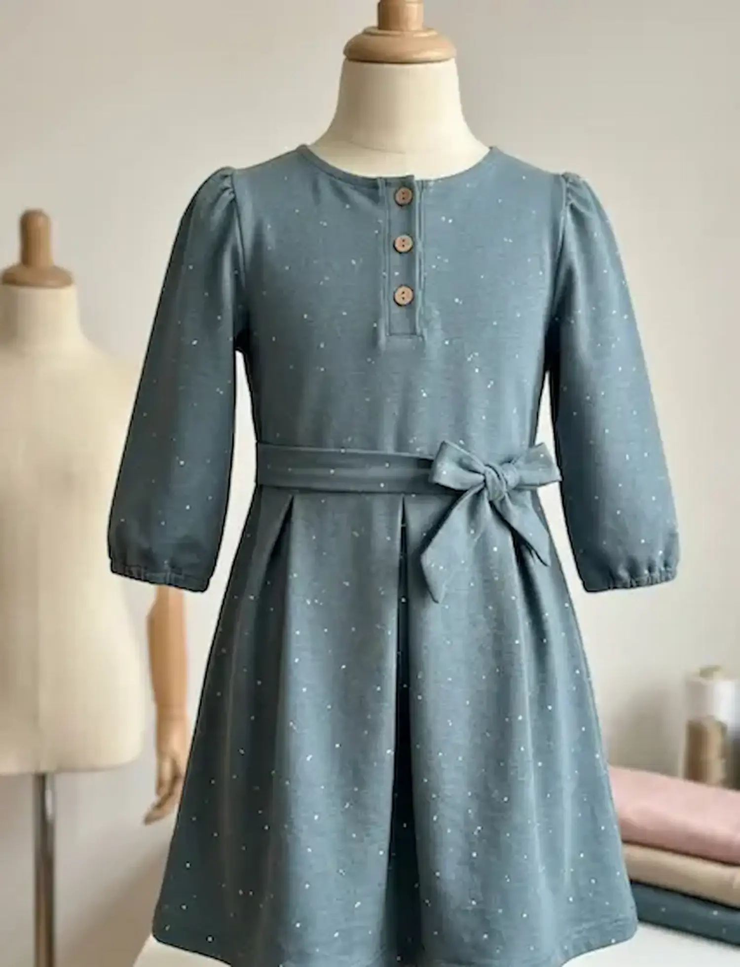 Genähtes Kinderkleid aus blaugrauem Jersey mit hellen Sprenkeln, Holzknöpfen und Taillenschleife auf einer Schneiderpuppe.