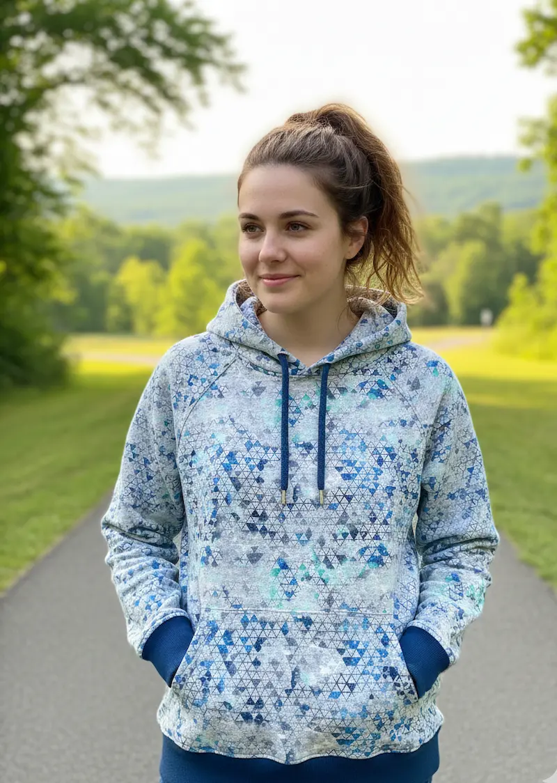 Eine Frau trägt einen Kapuzenpullover aus French Terry Meterware mit einem modernen geometrischen Dreiecksmuster in Blau- und Türkistönen vor einem ländlichen Hintergrund.
