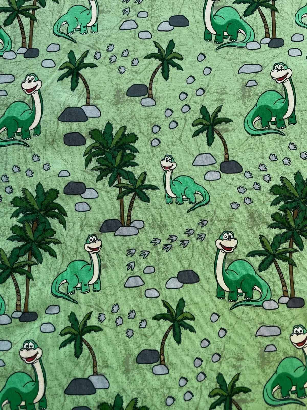 Hochwertiger Baumwolljersey als Meterware in Hellgrün mit einem fröhlichen Dinosaurier-Muster. Zu sehen sind freundliche, grüne Langhals-Dinos zwischen tropischen Palmen, grauen Steinen und kleinen Fußspuren im Allover-Design.