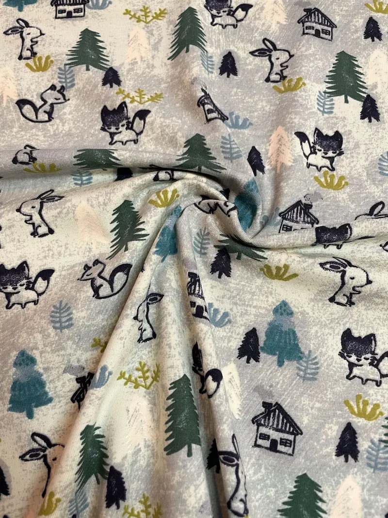 Ein weich drapierter Bio-Baumwolljersey-Stoff als Meterware in Hellgrau mit einem winterlichen Wald-Muster. Im Faltenwurf sind gezeichnete Füchse, Hasen, Tannenbäume und kleine Holzhütten zu sehen.