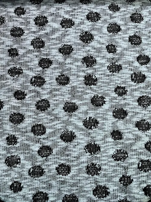 Weicher Strickstoff als Meterware in Hellgrau meliert, gemustert mit gleichmäßigen, eingestrickten schwarzen Punkten (Polka Dots). Die charakteristische, gemütliche Maschenstruktur des Feinstricks ist in der Nahaufnahme deutlich zu sehen.