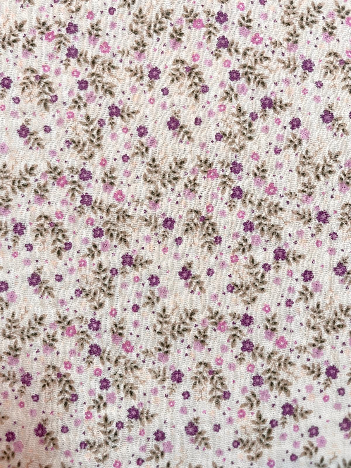 Nahaufnahme von weißem Musselin-Stoff mit kleinen lila Blumen und grünen Blättern, weiche Double-Gauze Struktur aus 100% Baumwolle.