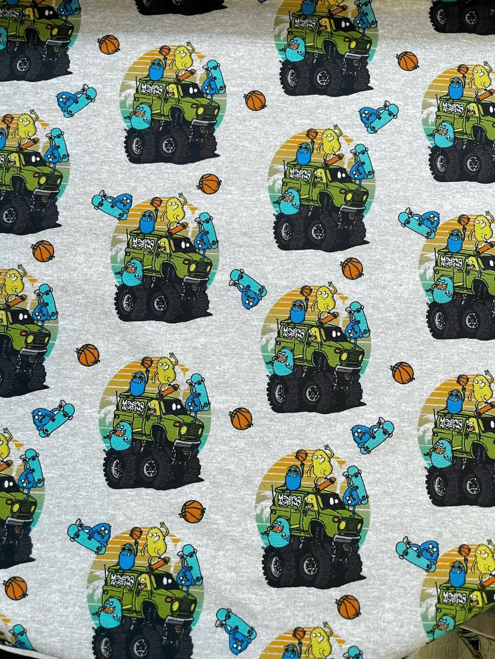 Grauer French Terry Stoff als Meterware bedruckt mit grünen Monster Trucks, Basketbällen und kleinen blauen sowie gelben Monstern.