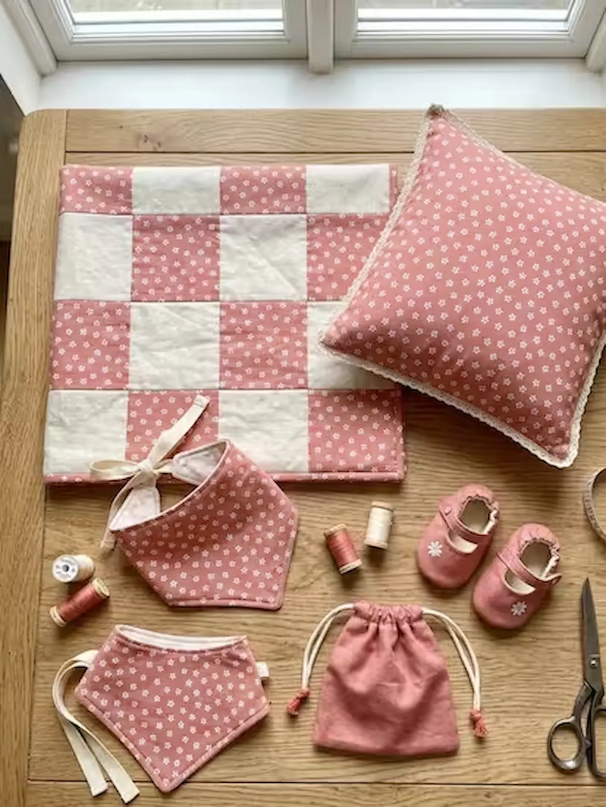 Nähbeispiele für Babys aus altrosafarbenem Baumwoll-Popeline-Stoff mit kleinen weißen Blumen: Eine Patchwork-Decke im Karomuster, ein Dekokissen mit Spitzenborte, zwei Halstücher (Sabberlatz), ein kleiner Beutel und passende Babyschuhe auf einem Holztisch
