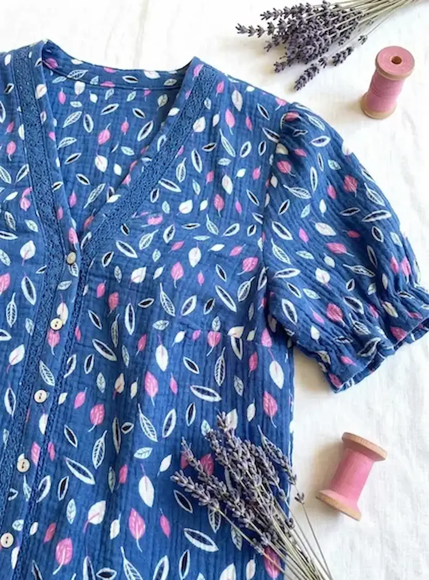 Dunkelblaue Bluse aus Musselin-Stoff mit rosa und weißen Blättern, dekoriert mit Lavendel und Garnrollen auf hellem Untergrund.