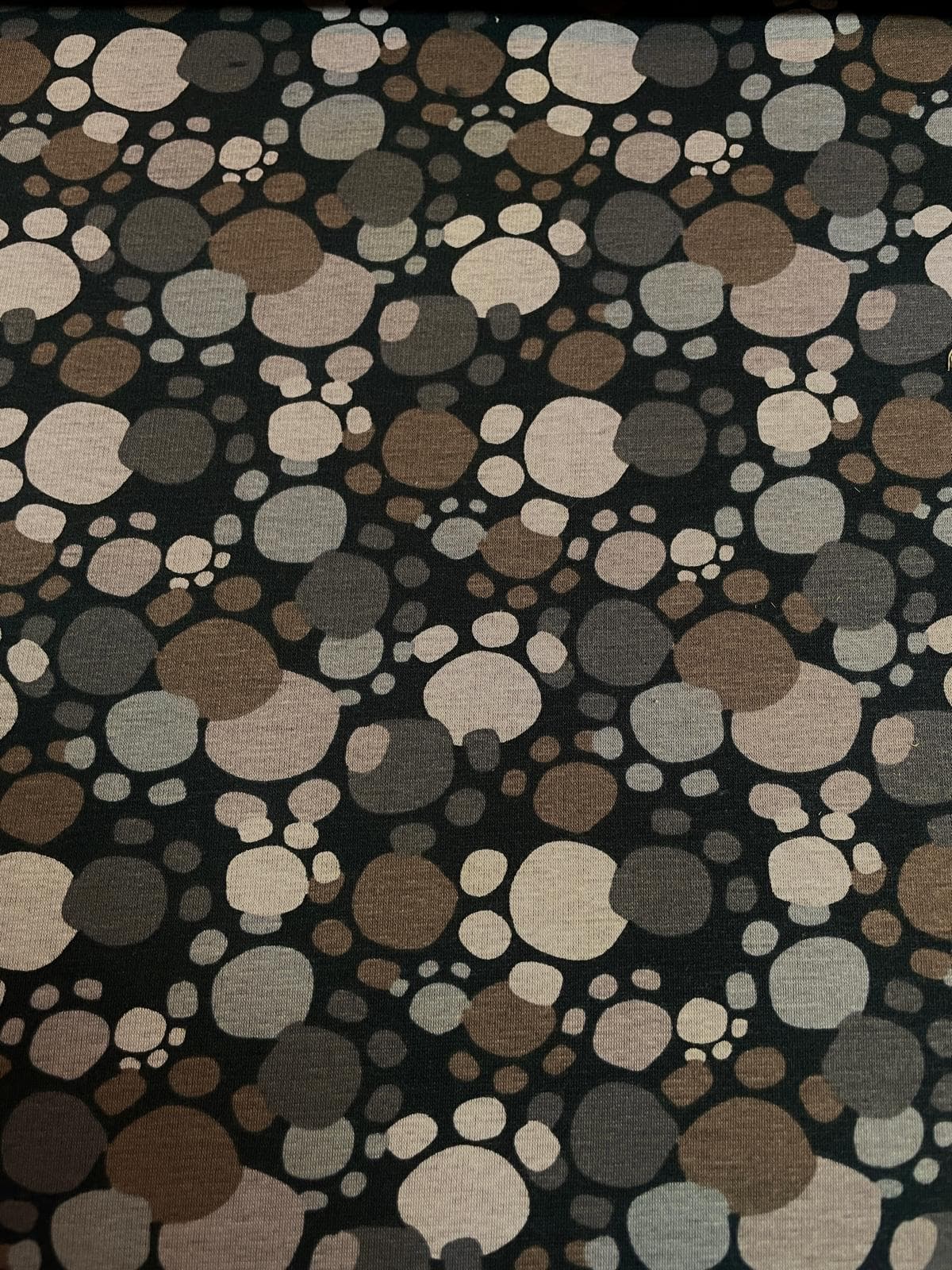 Dunkler Alpenfleece-Stoff mit einem Muster aus abstrakten Pfotenabdrücken und Punkten in verschiedenen Erdtönen wie Braun, Taupe, Beige und Grau auf schwarzem Hintergrund.