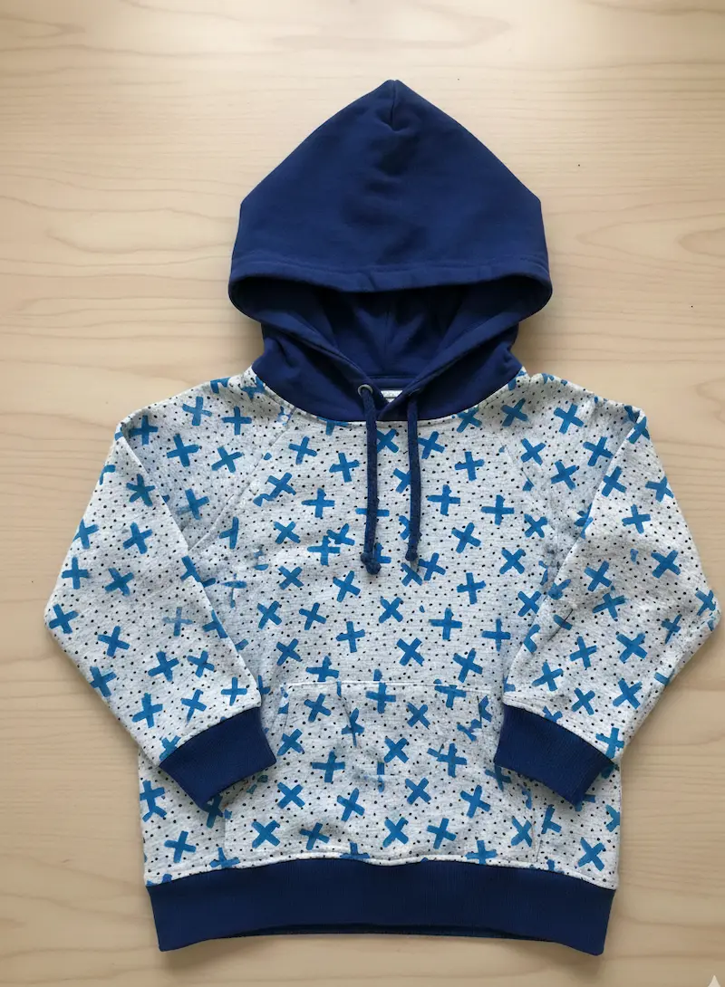 Genähter Raglan-Kinderhoodie aus grau-meliertem French Terry mit blauen Kreuzen und schwarzen Punkten, kombiniert mit einer Kapuze und Bündchen in Dunkelblau