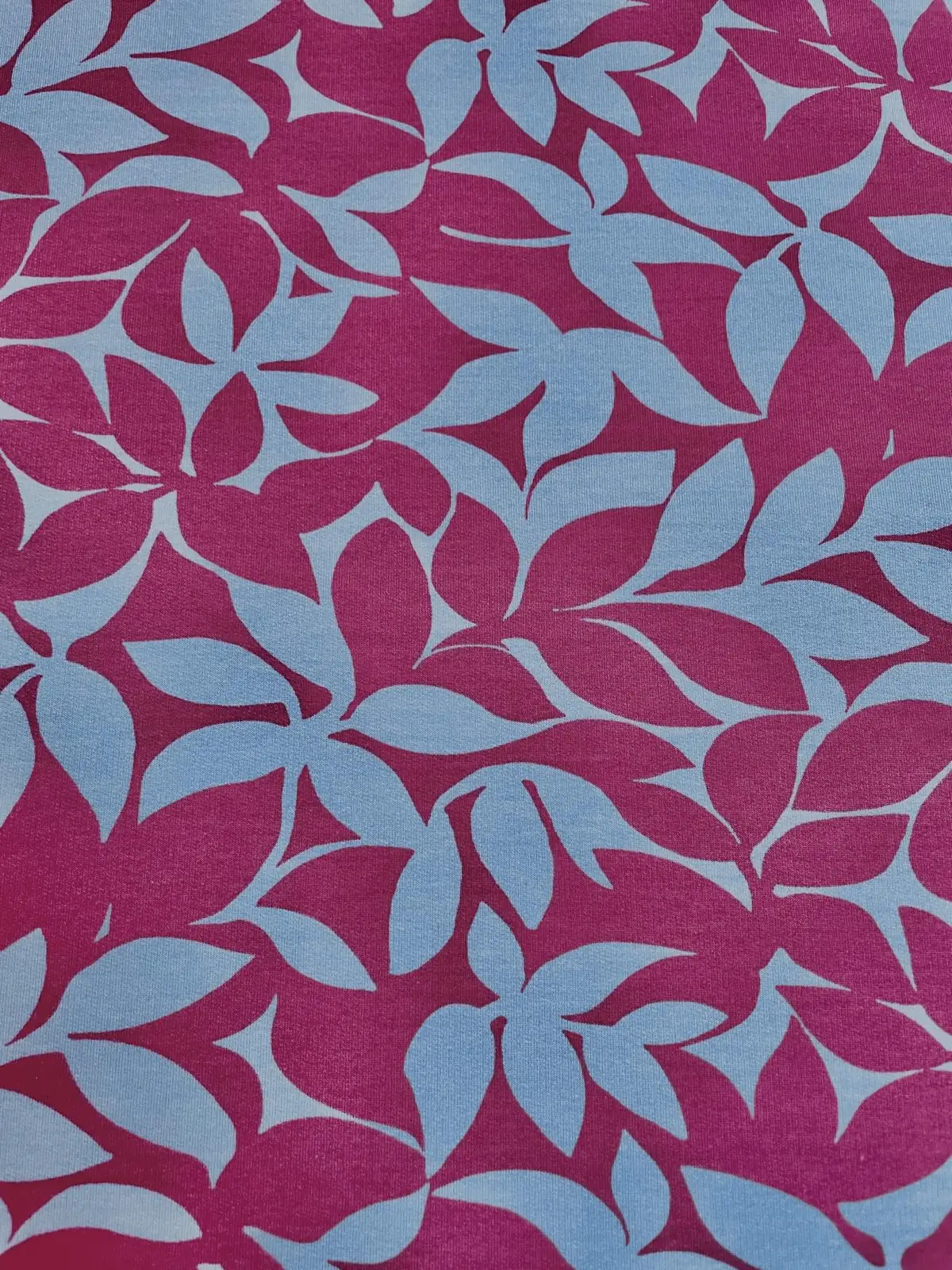 Weicher Sweatstoff mit einem großflächigen, abstrakten Blumenmuster in hellem Blau auf einem kräftigen beerenfarbenen Hintergrund. Der Stoff ist spiralförmig drapiert und zeigt seine geschmeidige Textur.