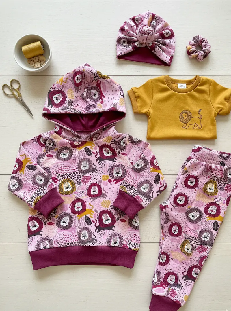 Designbeispiel für French Terry Meterware: Ein selbstgenähter Kinder-Hoodie mit Kapuze und passende Sweathose aus pinkem Löwen-Stoff, kombiniert mit einem senfgelben T-Shirt, einem Turban-Haarband und Nähzubehör auf hellem Holzboden.